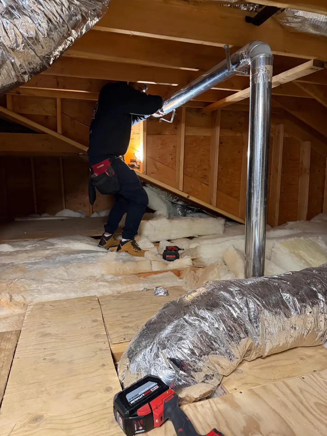 A technician in an attic beside a rigid metal vent pipe, with flexible insulated ducts on the floor.