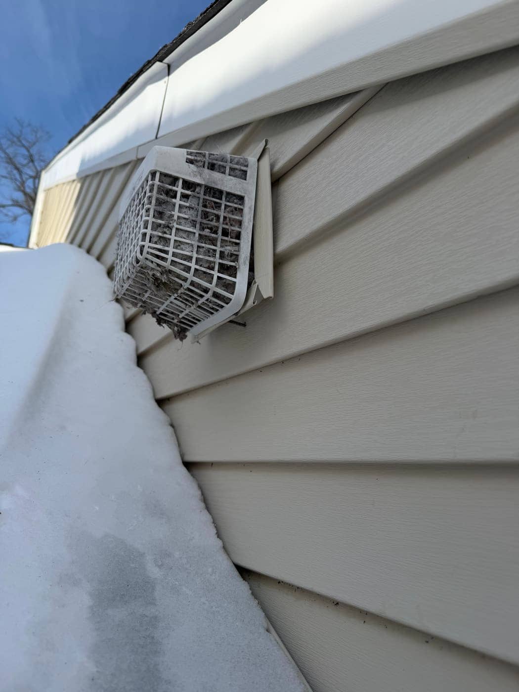 A dryer vent hood on vinyl siding packed with lint, with snow piled on the ground below.