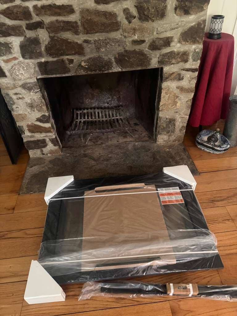 An open fieldstone fireplace with a grate inside and a boxed glass door kit on the floor in front of it.