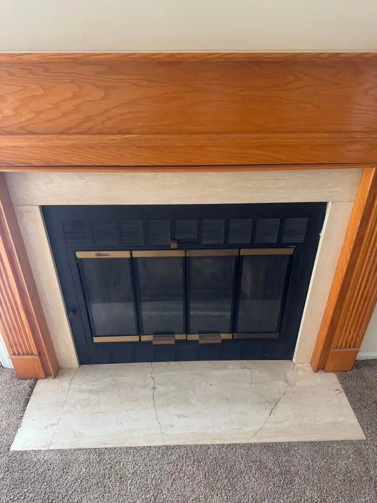 A fireplace with black glass doors and brass-colored trim under an oak mantel, with a pale marble hearth.