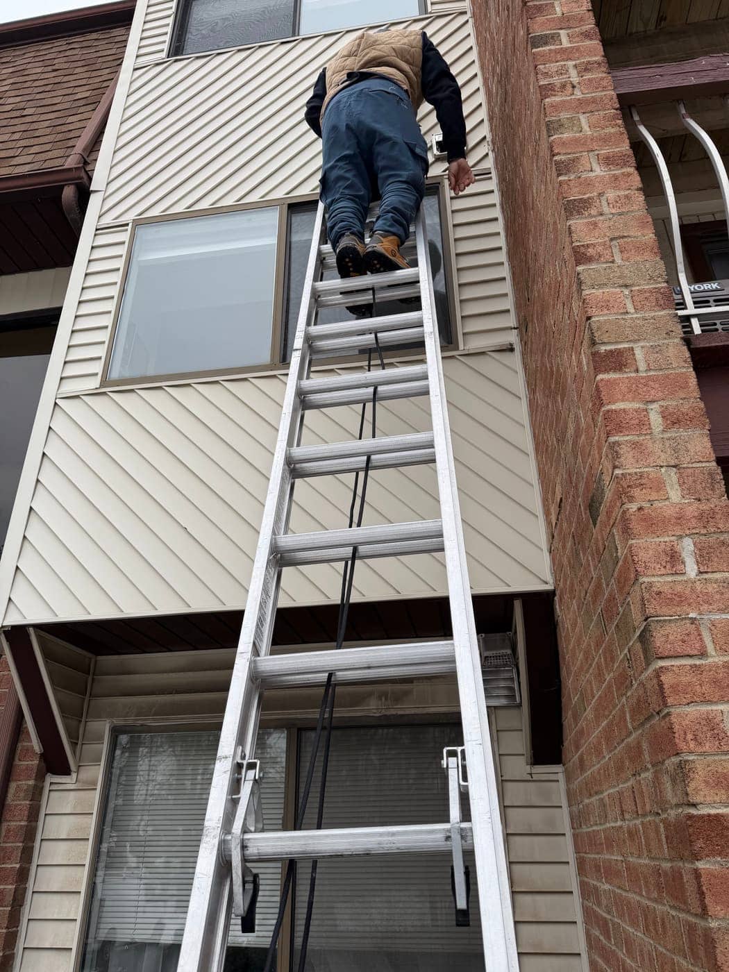 A technician climbing an extension ladder against a building with tan siding and a brick wall.