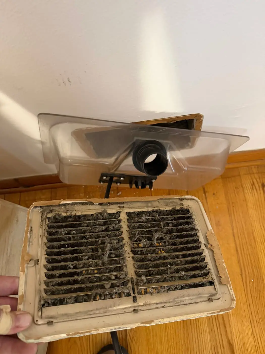 A register grille clogged with dust and lint, held up next to a floor opening fitted with a vacuum shroud.