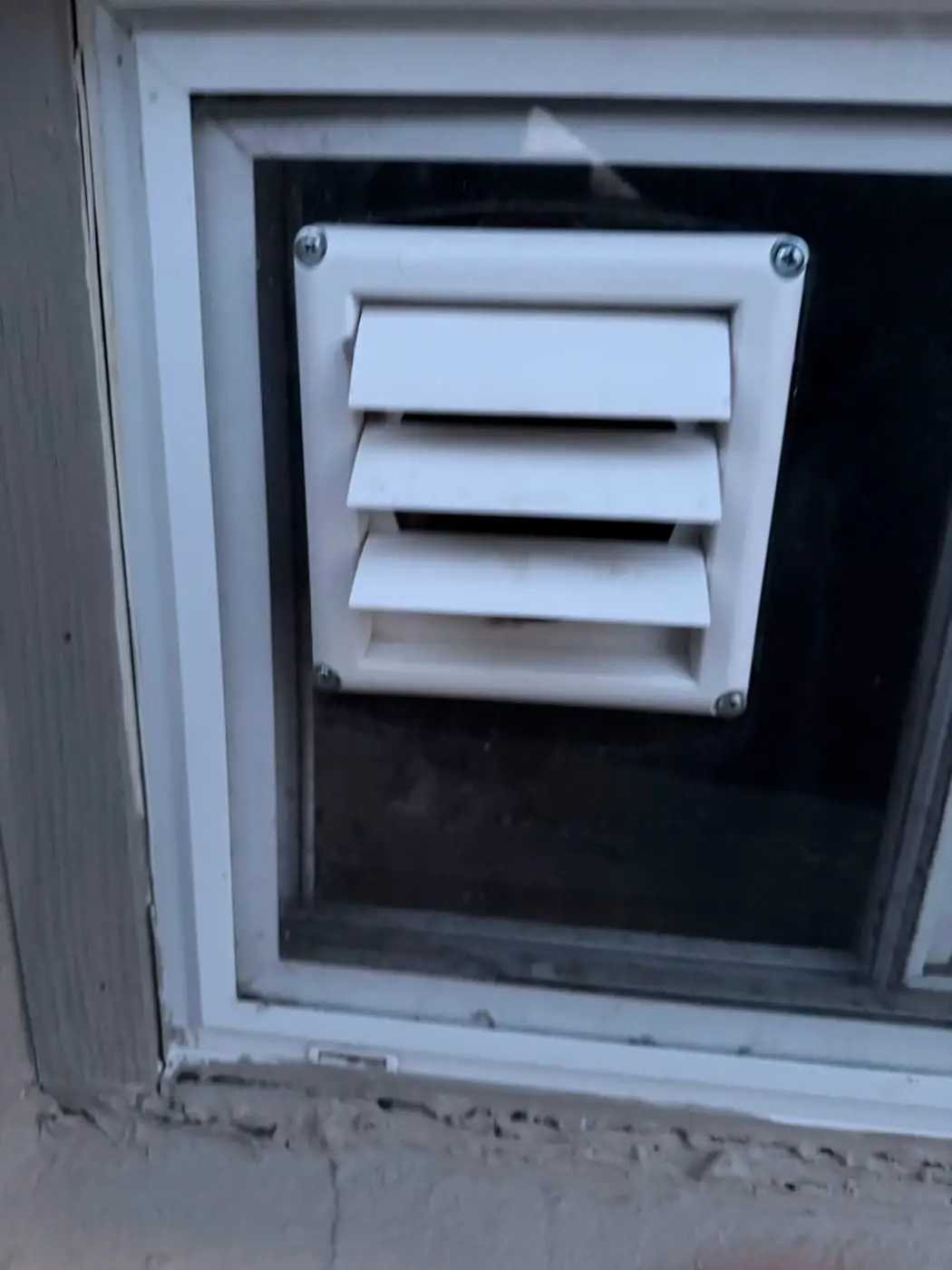 A white louvered vent hood mounted in a panel that fills a basement window.