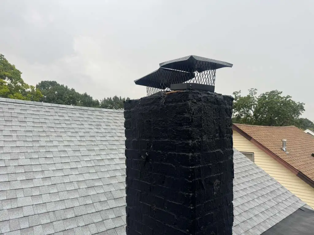 A mesh chimney cap on a brick chimney coated black, rising through a gray shingle roof.