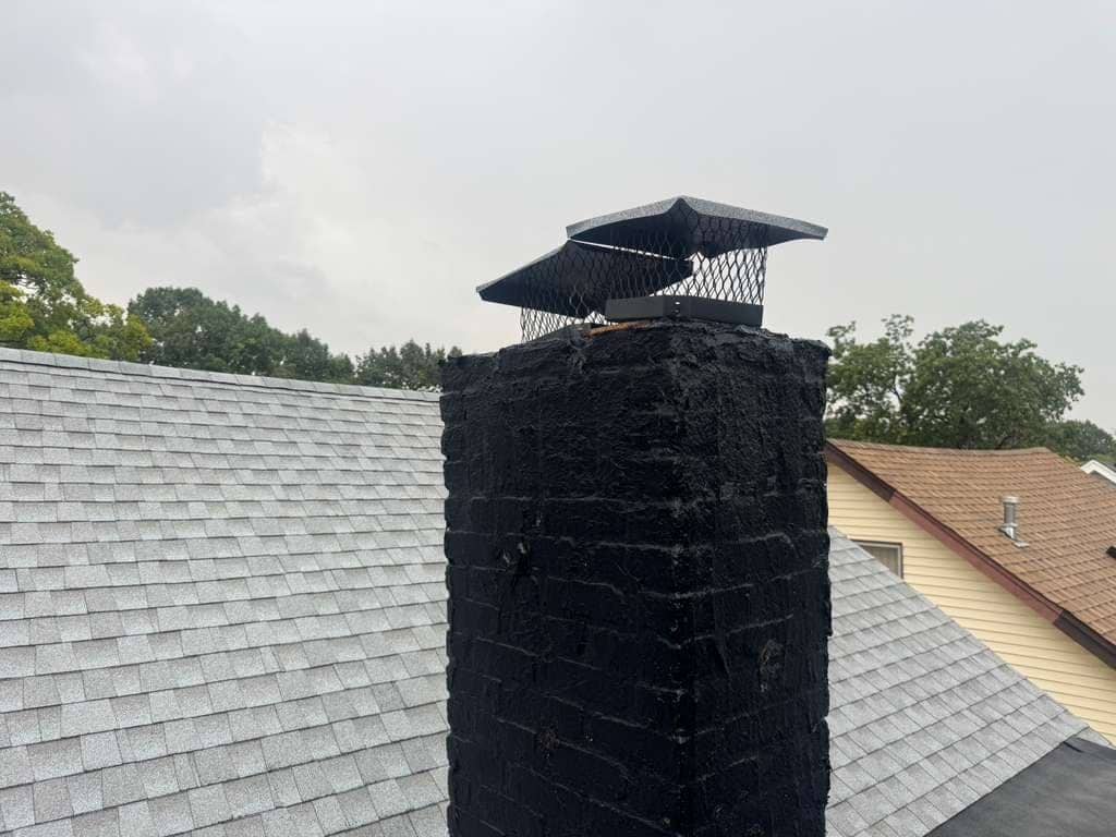 A mesh chimney cap on a brick chimney coated black, rising through a gray shingle roof.
